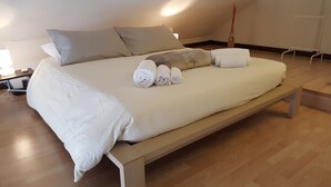 Ein ordentlich bezogenes Bett mit aufgerollten Handtüchern, eine Holzbank und ein Nachttisch mit einer Lampe.
