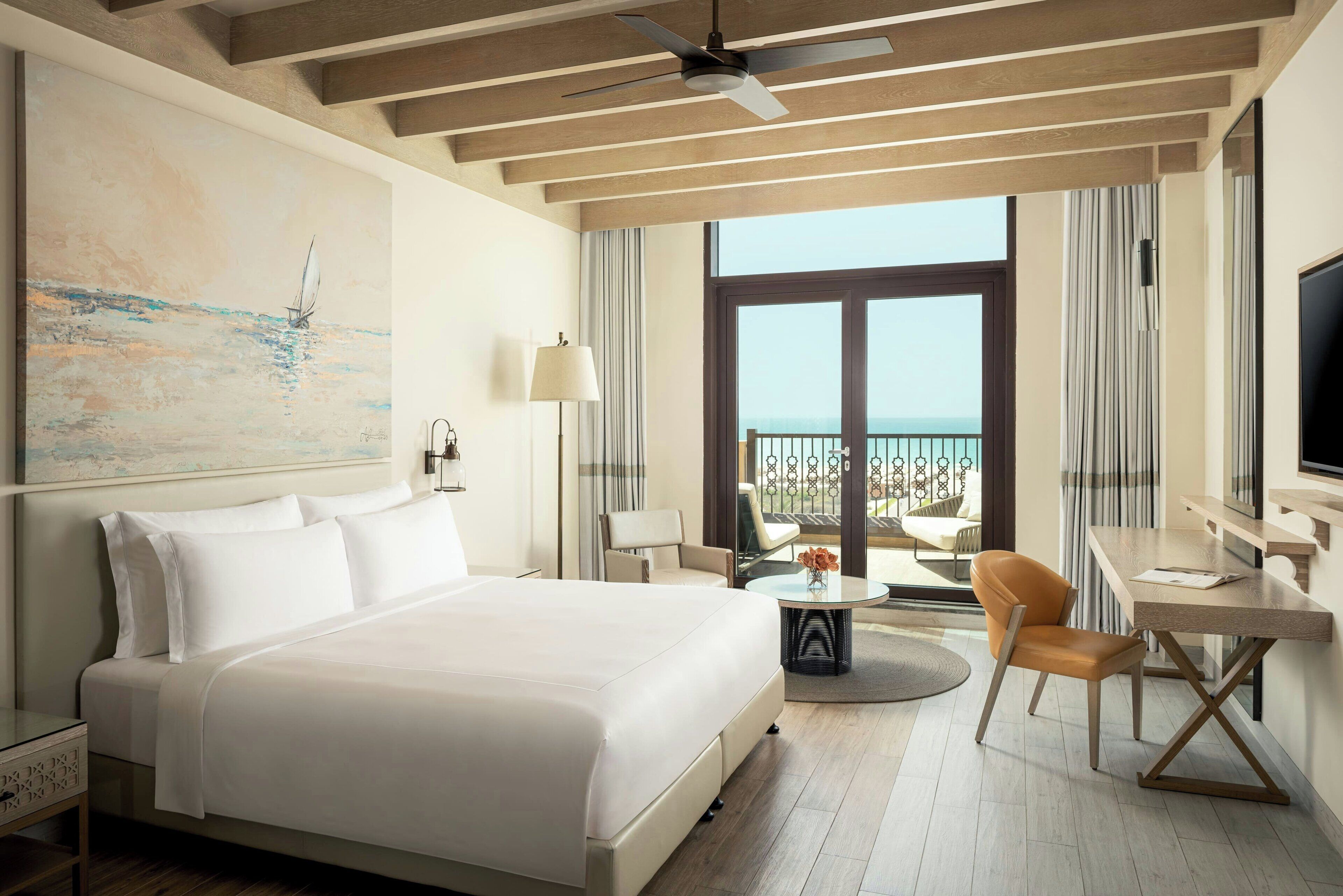 A hotel room with a bed, a desk, a chair, a TV, a painting, and a balcony with a view.