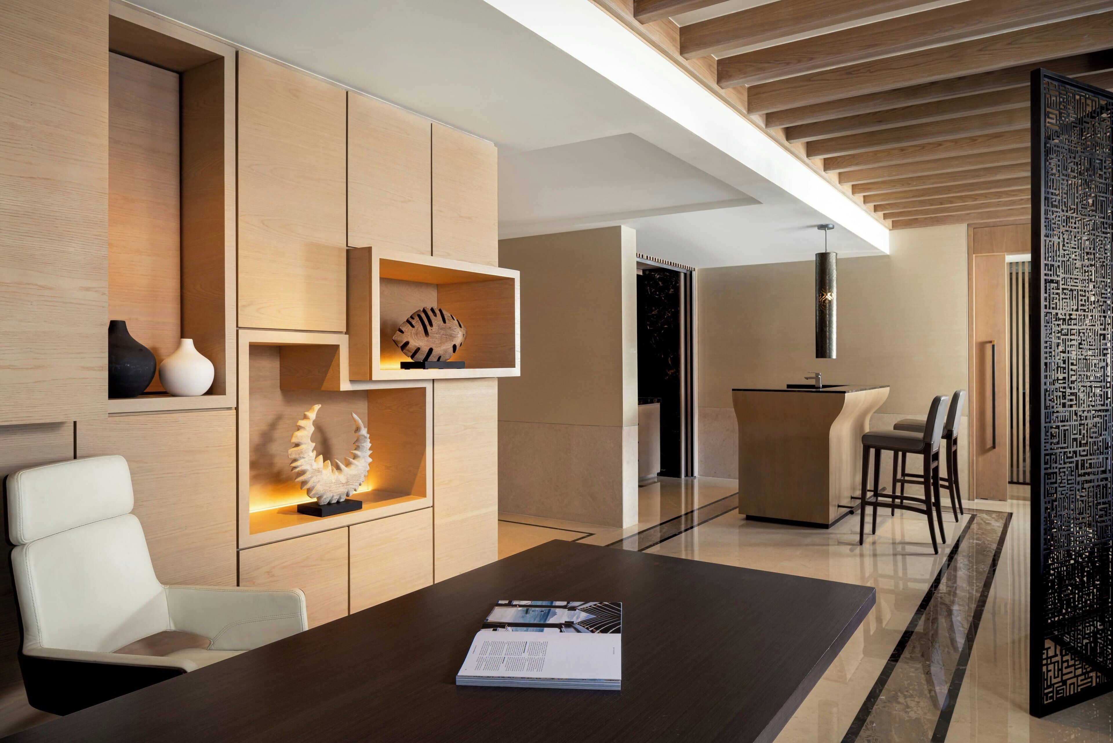 A modern interior with a wooden desk, a white chair, and a dining area with a kitchen in the background.
