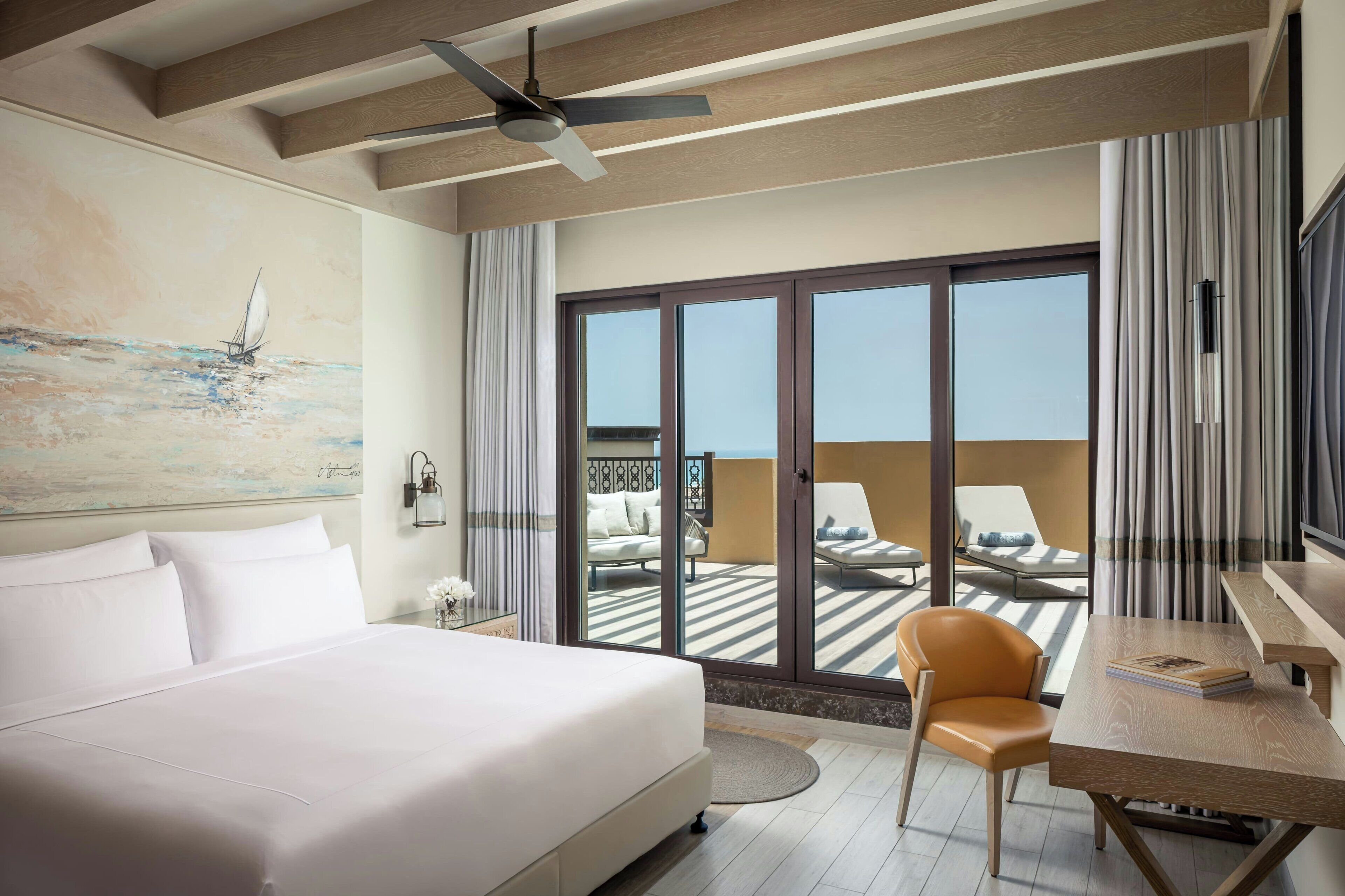Une chambre d’hôtel comprenant un lit, un bureau, une chaise, un ventilateur de plafond et une vue sur le balcon, qui est aménagé avec du mobilier d’extérieur.