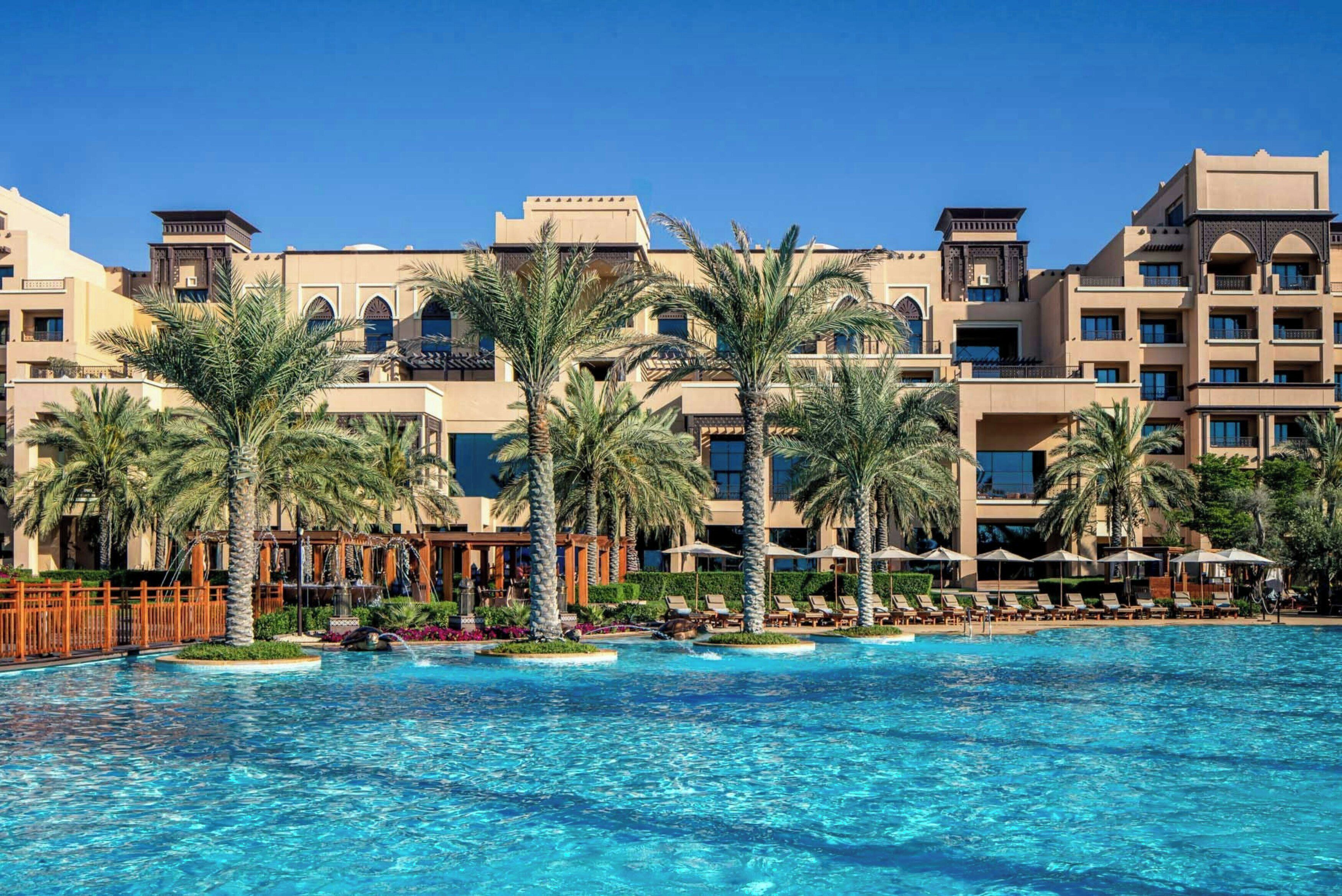 A hotel with a swimming pool, palm trees, and a clear blue sky.