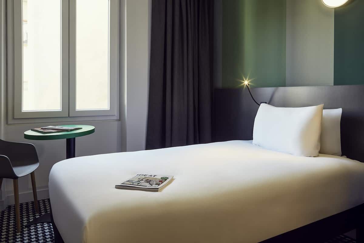 Ein Hotelzimmer mit Bett, einem kleinen Tisch mit einer Zeitschrift, einem Sessel, einem Fenster mit Vorhängen und einer Wandleuchte.