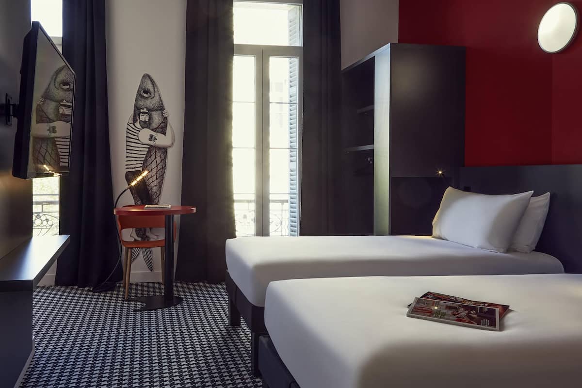 A hotel room with two beds, a desk with a chair, a large mirror, and a window with curtains.