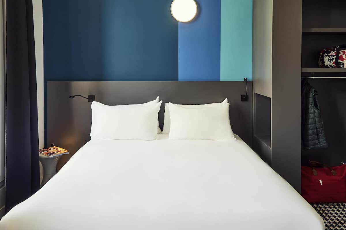 A hotel room with a bed, a nightstand, a lamp, a wardrobe, and a red bag.