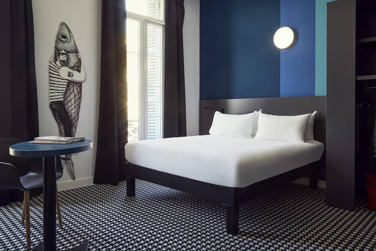 Ein Hotelzimmer mit einem Bett, einem runden Tisch, einem Stuhl, einer Wanduhr und einem Fenster mit Vorhängen.