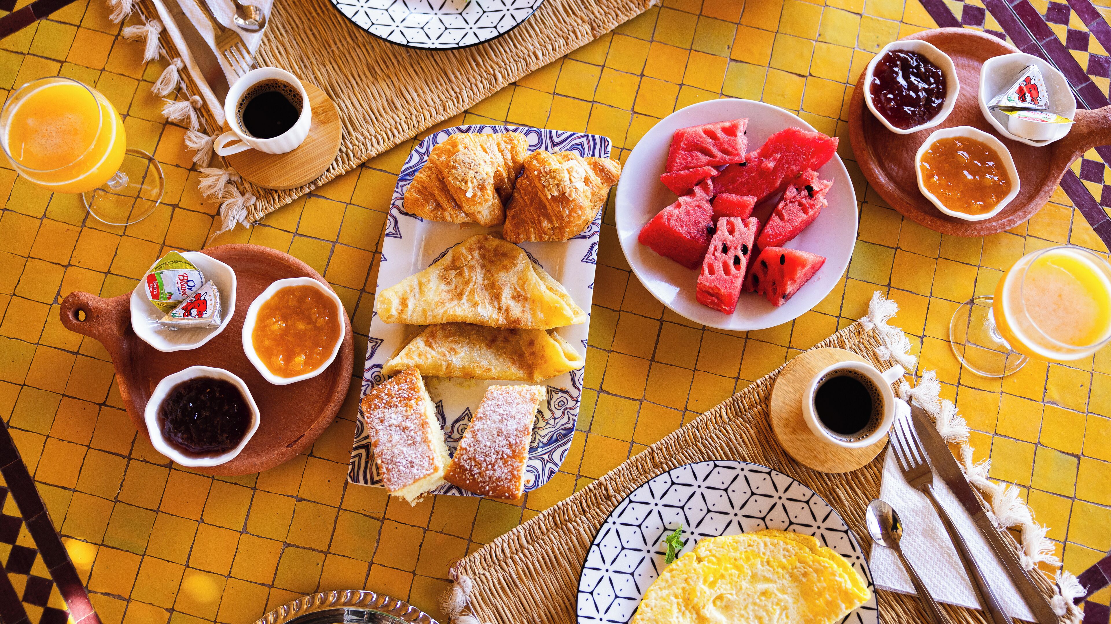A breakfast setting with croissants, watermelon, and various jams on a yellow-tiled table.