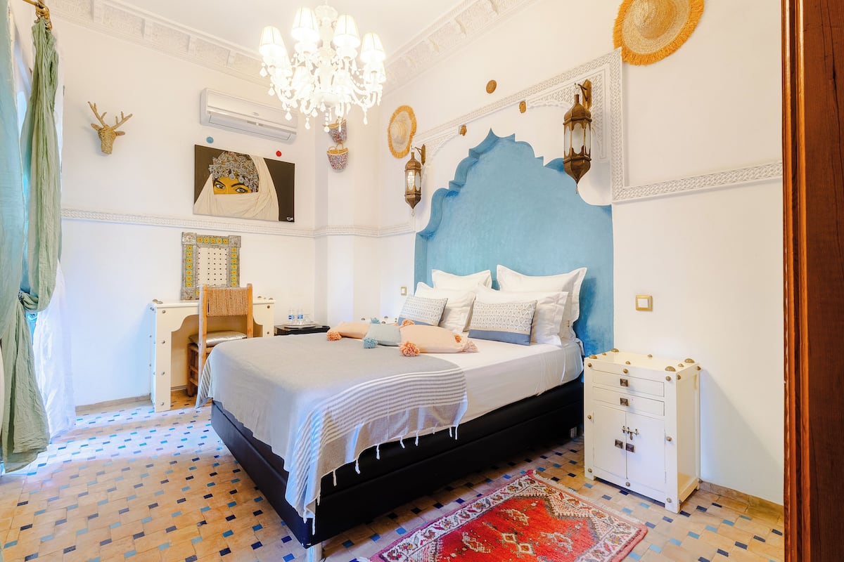 Une chambre à coucher avec un grand lit, une table de chevet, une commode, un lustre et des œuvres d’art décoratives au mur.