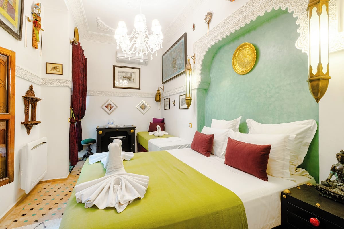 Une chambre d’hôtel comprenant un lit, un bureau, une chaise, un radiateur, un lustre et un mur agrémenté d’éléments décoratifs.