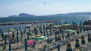 Uma praia com fileiras de espreguiçadeiras e guarda-sóis, um molhe rochoso e uma vista clara do mar e colinas ao longe.