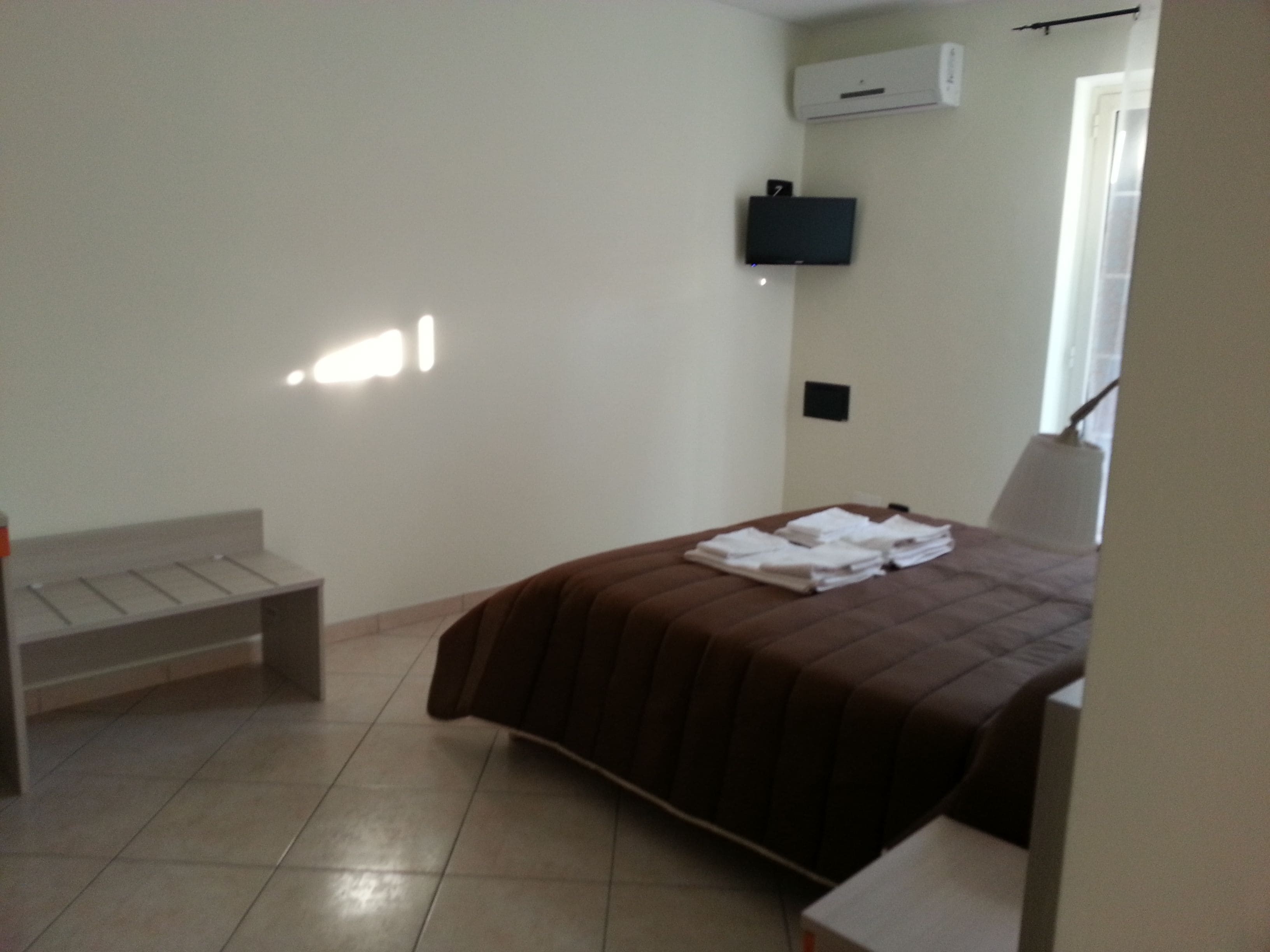 Un dormitorio con cama, un banco, un televisor montado en la pared y un equipo de aire acondicionado.