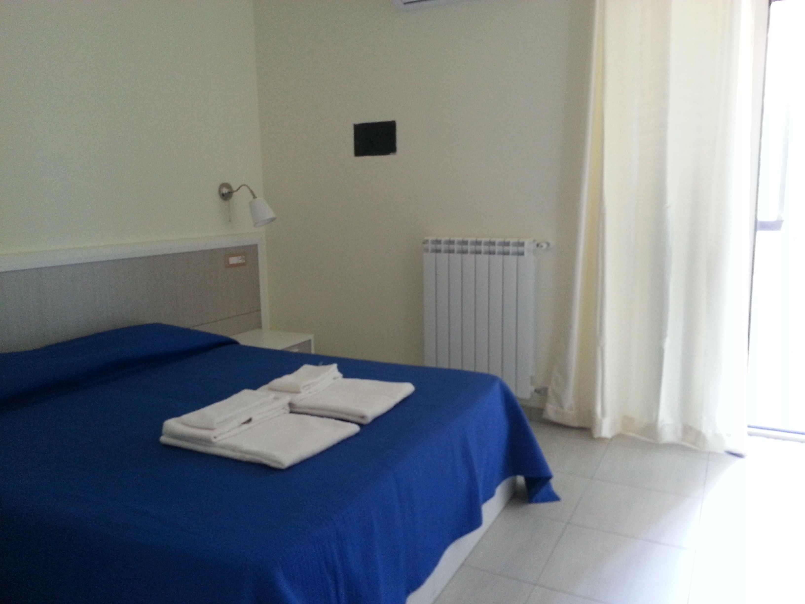 A hotel room with a bed, a nightstand, a lamp, a radiator, and a window with curtains.
