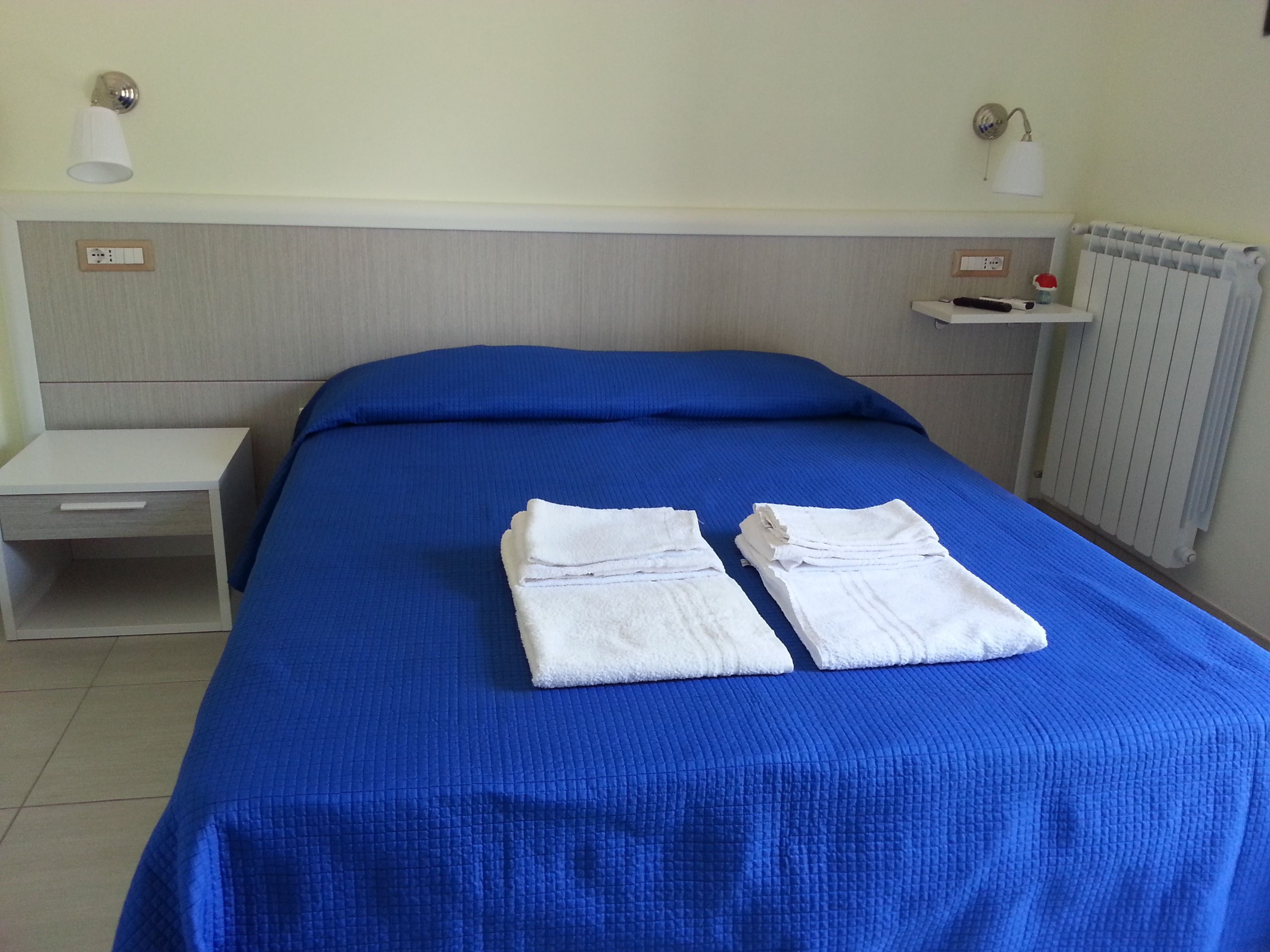 A neatly made bed with blue bedding and white towels, a bedside table, a nightstand, and a wall-mounted lamp.