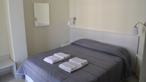 Una cama bien hecha con ropa de cama gris y toallas blancas, una mesita de noche, un espejo fijado a la pared y dos lámparas de pared.