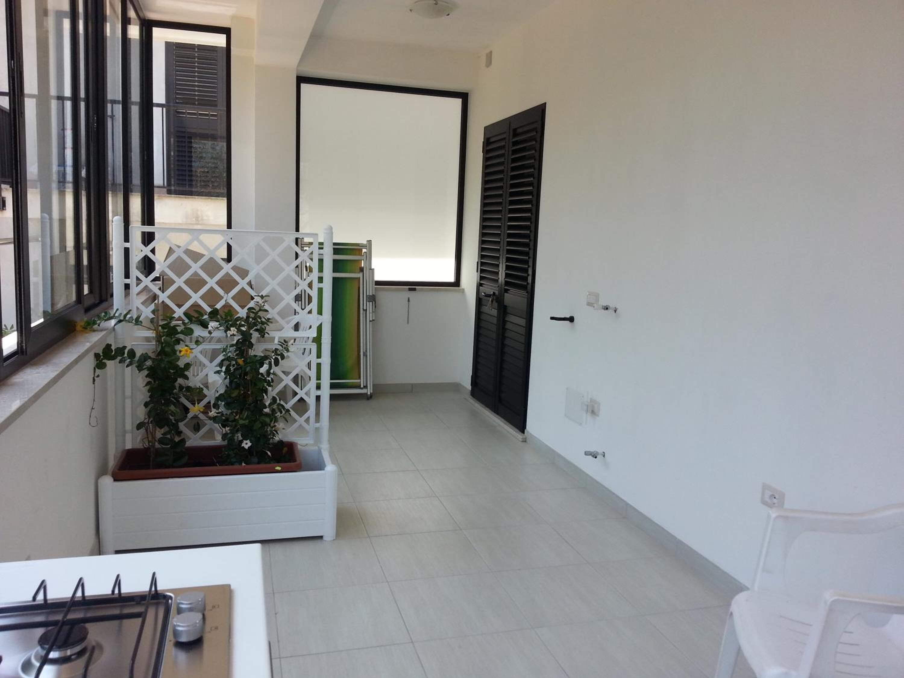 Una habitación pequeña y limpia con piso de baldosas blancas, un escritorio blanco con estufa a gas, una silla blanca, un panel blanco con una planta y una puerta con persiana negra.