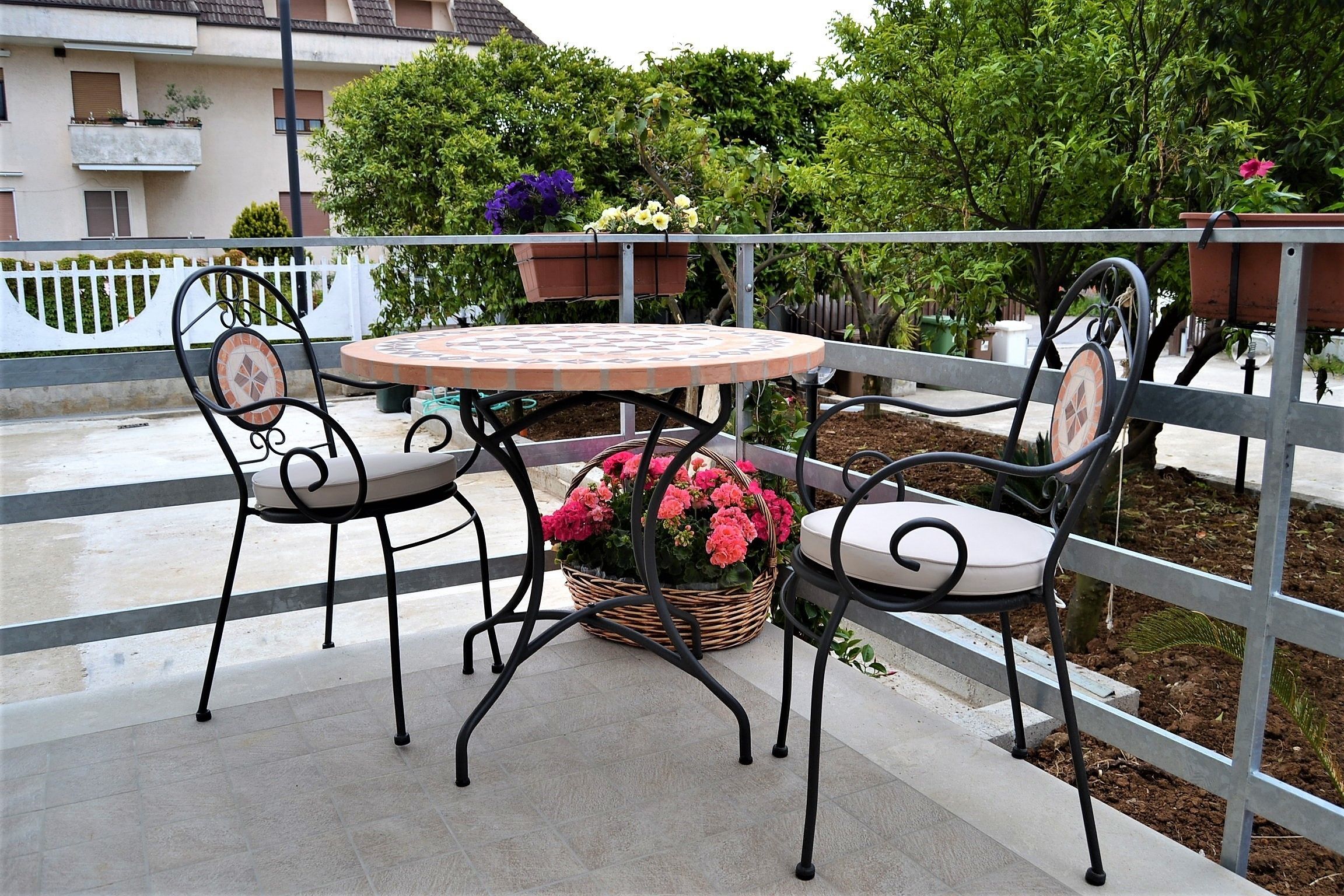 En udendørs terrasse med et rundt bord og fire stole, omgivet af et metalhegn.
