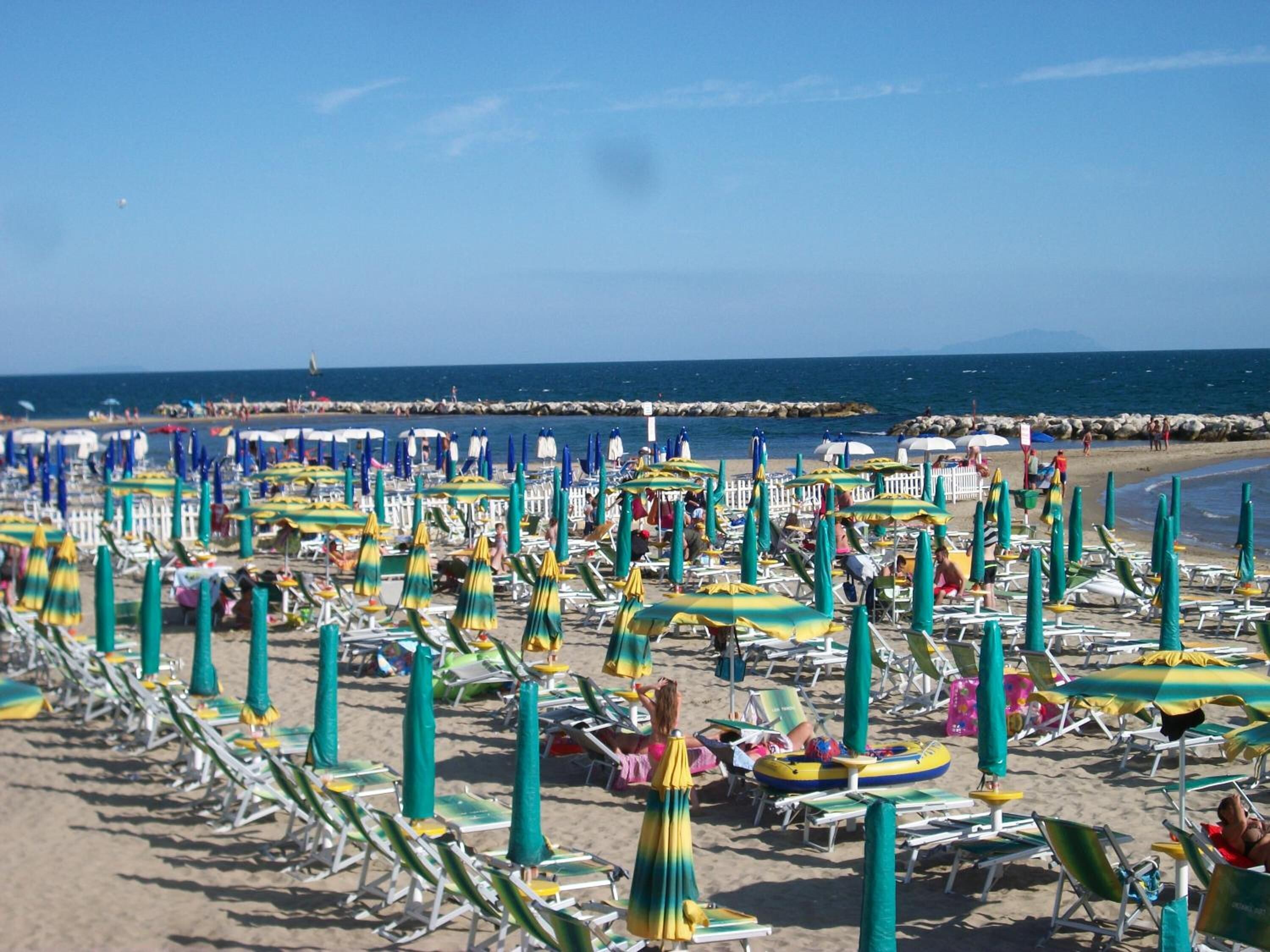 Une plage bordée de rangées de chaises longues et de parasols, une digue rocheuse en arrière-plan et l’océan.