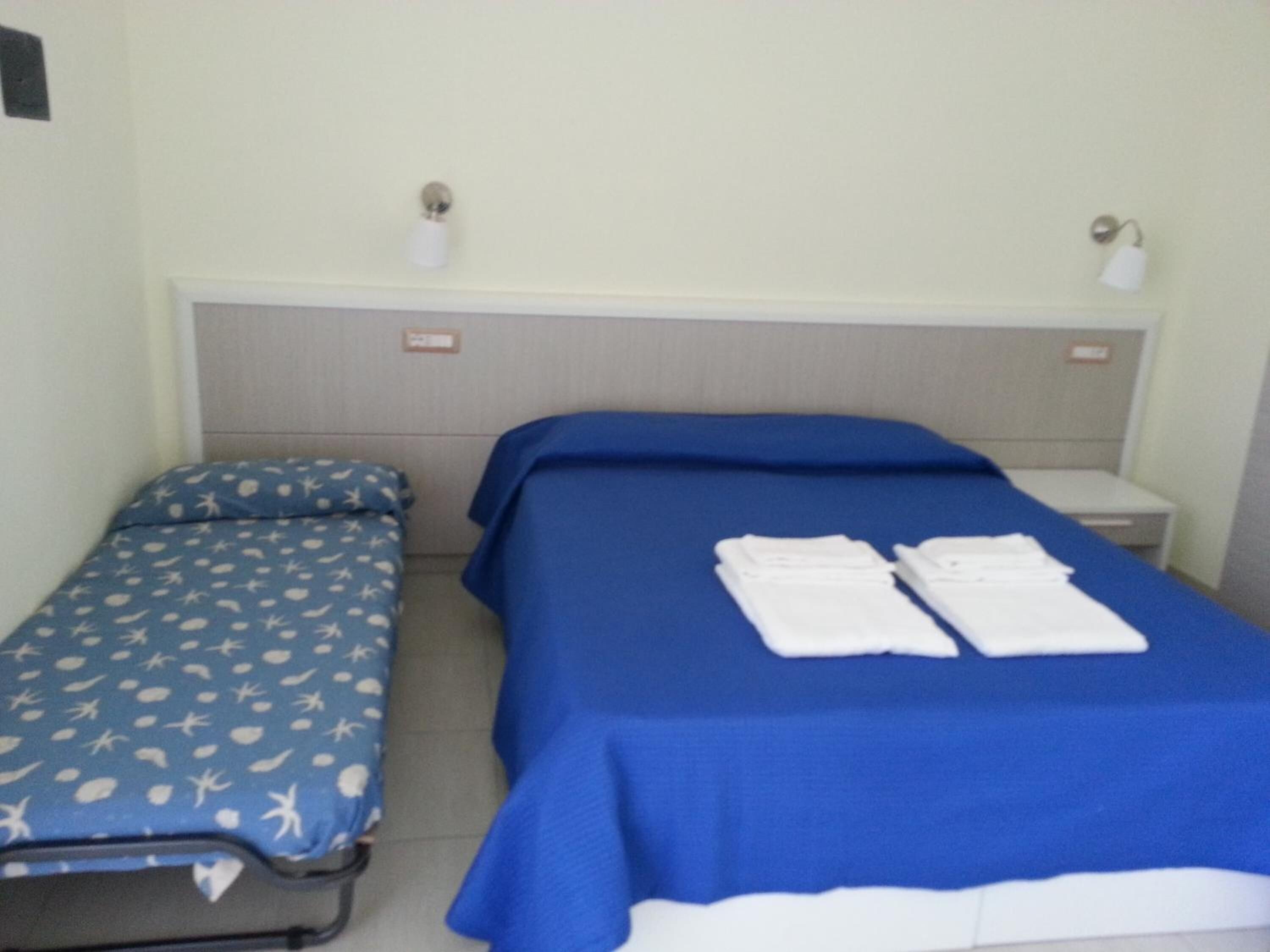 A room with two beds, one with a blue bedspread and the other with a blue bedspread with a white pattern. There are two white towels on the larger bed.