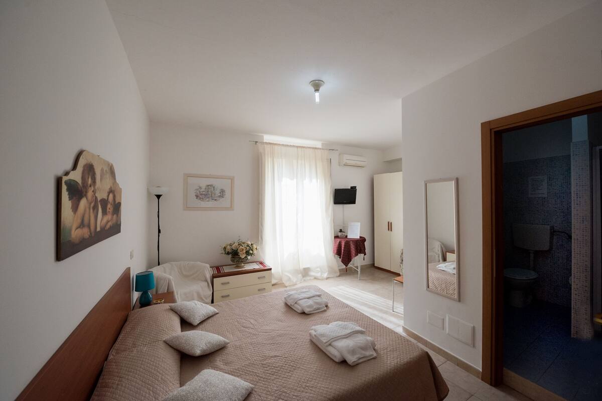 A bedroom with a bed, a bedside table, a lamp, a mirror, and a bathroom visible through an open door.