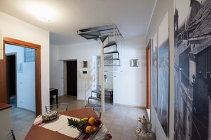 Un coin repas avec une table dressée pour deux personnes, un panier de fruits et un objet décoratif. Un escalier en colimaçon est visible au fond.