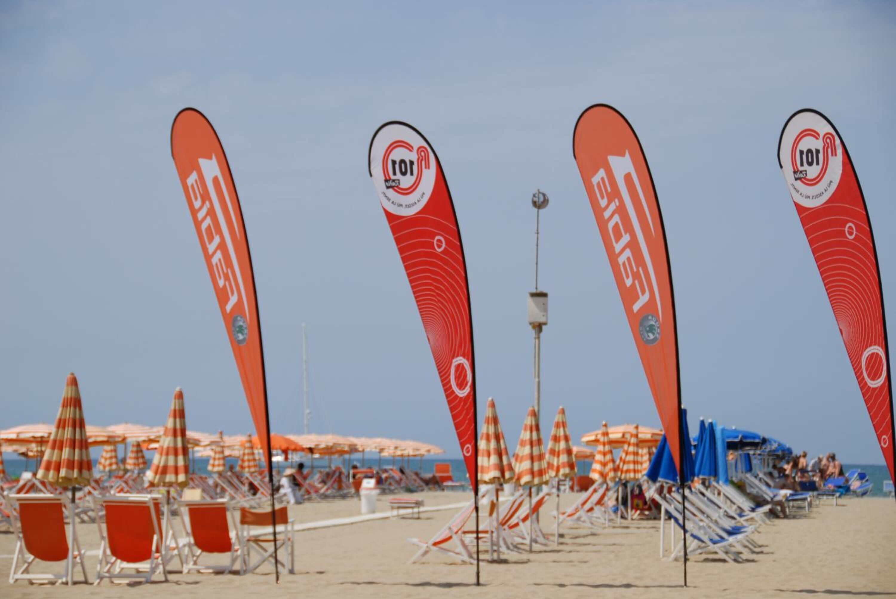 A beach with rows of sun loungers and umbrellas, and red flags with logos.