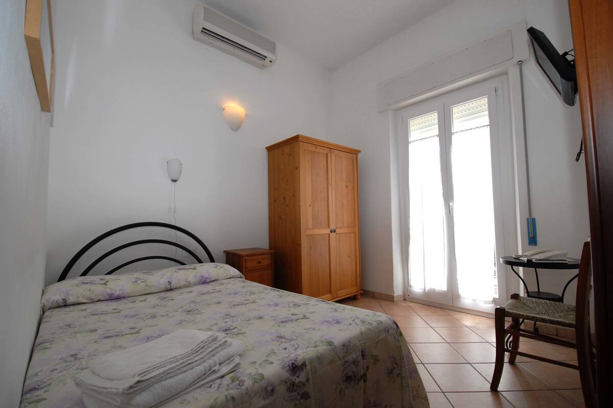 A bedroom with a bed, a wooden wardrobe, a desk with a chair, and a window with blinds.