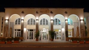 Un edificio con una serie de arcos y columnas, un gran acceso con puertas de cristal y plantas en macetas en la entrada.
