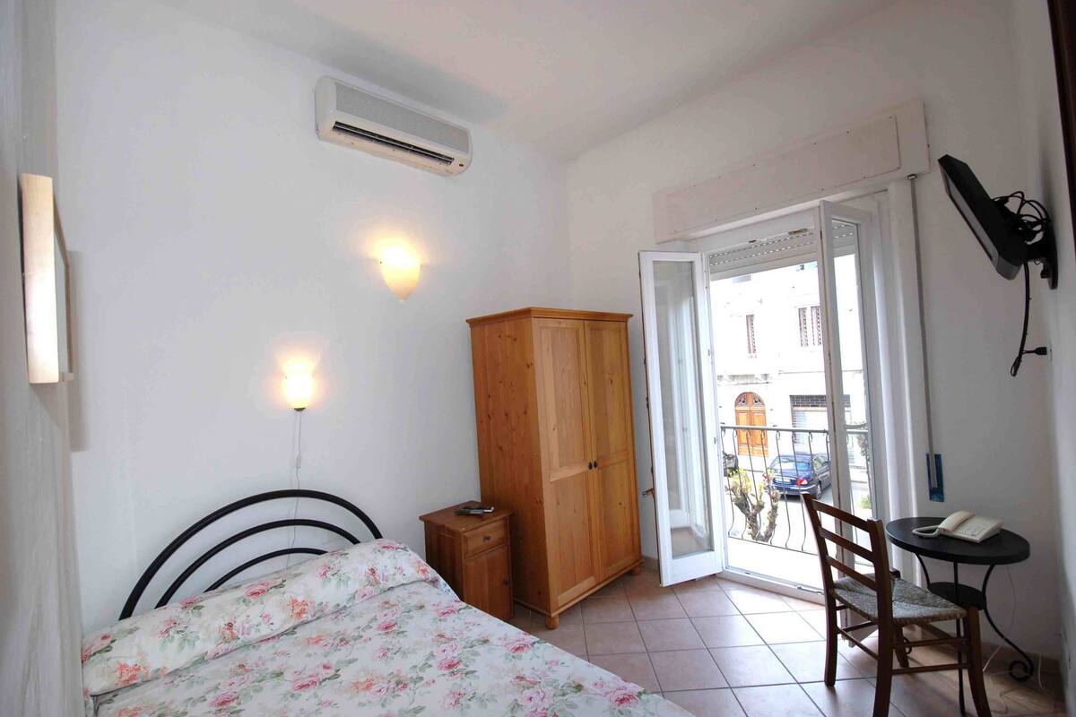 A bedroom with a bed, a wooden wardrobe, a desk with a chair, and a balcony with a view of the street.