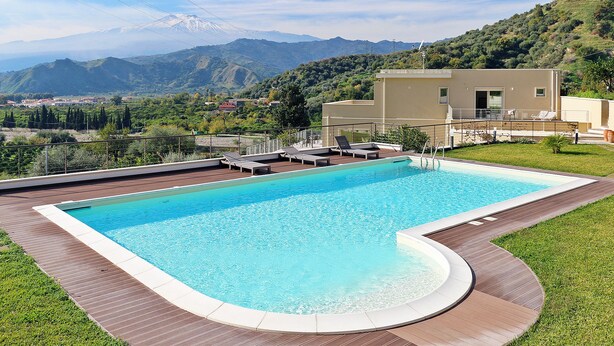 Una piscina rectangular con una terraza de madera y un borde curvo, rodeada de césped y un edificio con balcón.
