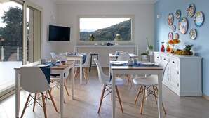Comedor moderno con mobiliario blanco, un ventanal con vistas a colinas y una pared decorada con platos.