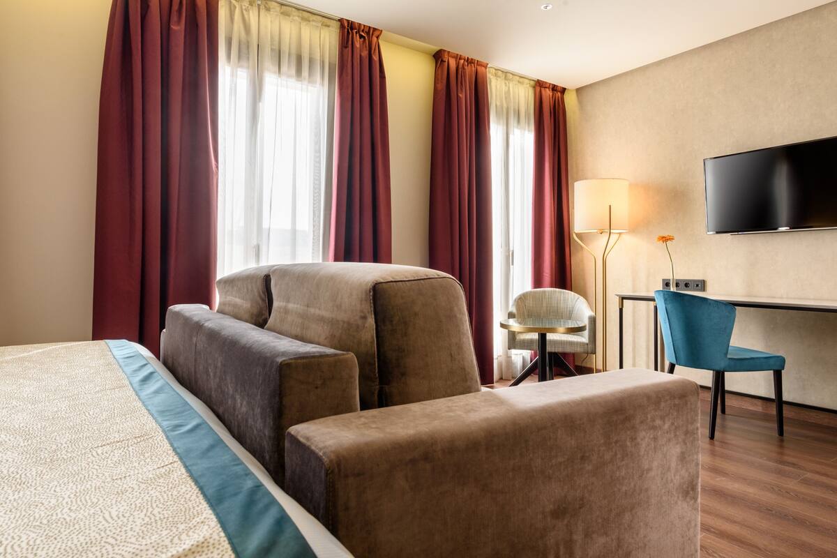 Een hotelkamer met een groot bed, twee fauteuils, een bureau met stoel, een televisie en ramen met gordijnen.