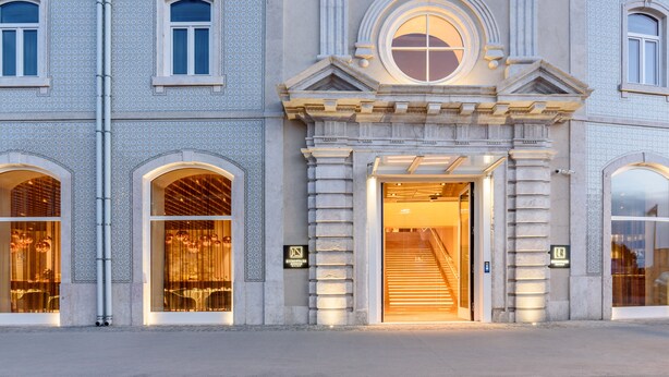 Un imposant bâtiment doté de grandes fenêtres en arc de cercle, d’une entrée centrale avec un escalier et d’une fenêtre circulaire décorative au-dessus de la porte.