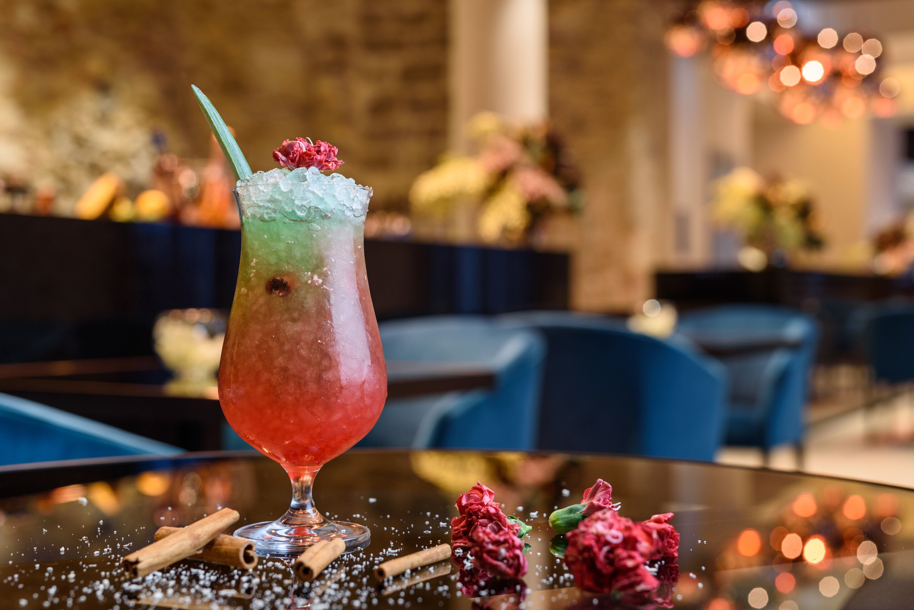 A cocktail glass with a gradient color effect, garnished with a flower and a straw, placed on a table with scattered spices and flowers.