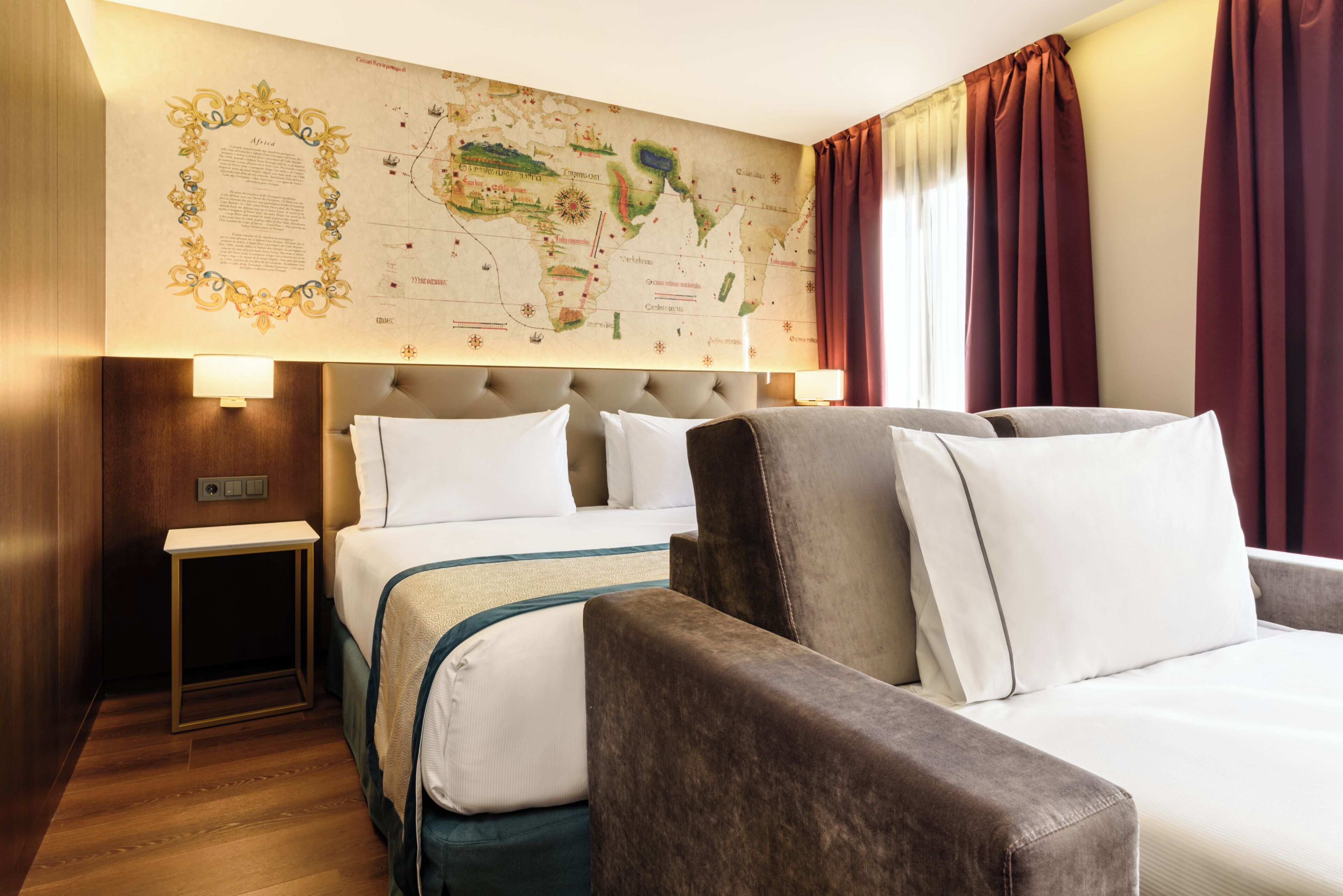 Habitación de hotel con una cama grande, un sofá, una mesita de noche, un mapa en la pared y una ventana con cortinas.