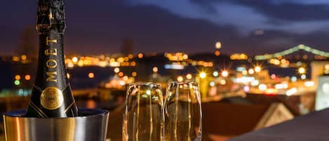 Una botella de champán Moët en un cubo metálico con dos copas de champán vacías sobre una mesa, con un paisaje urbano de noche al fondo.