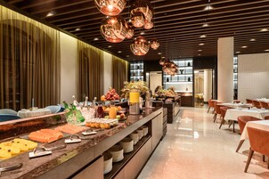 Un'area buffet con una varietà di cibi, tra cui frutta, verdura e bevande. Tavoli e sedie disposti per consumare i pasti, e l'ambiente presenta un design moderno con pannelli in legno e illuminazione a sospensione.