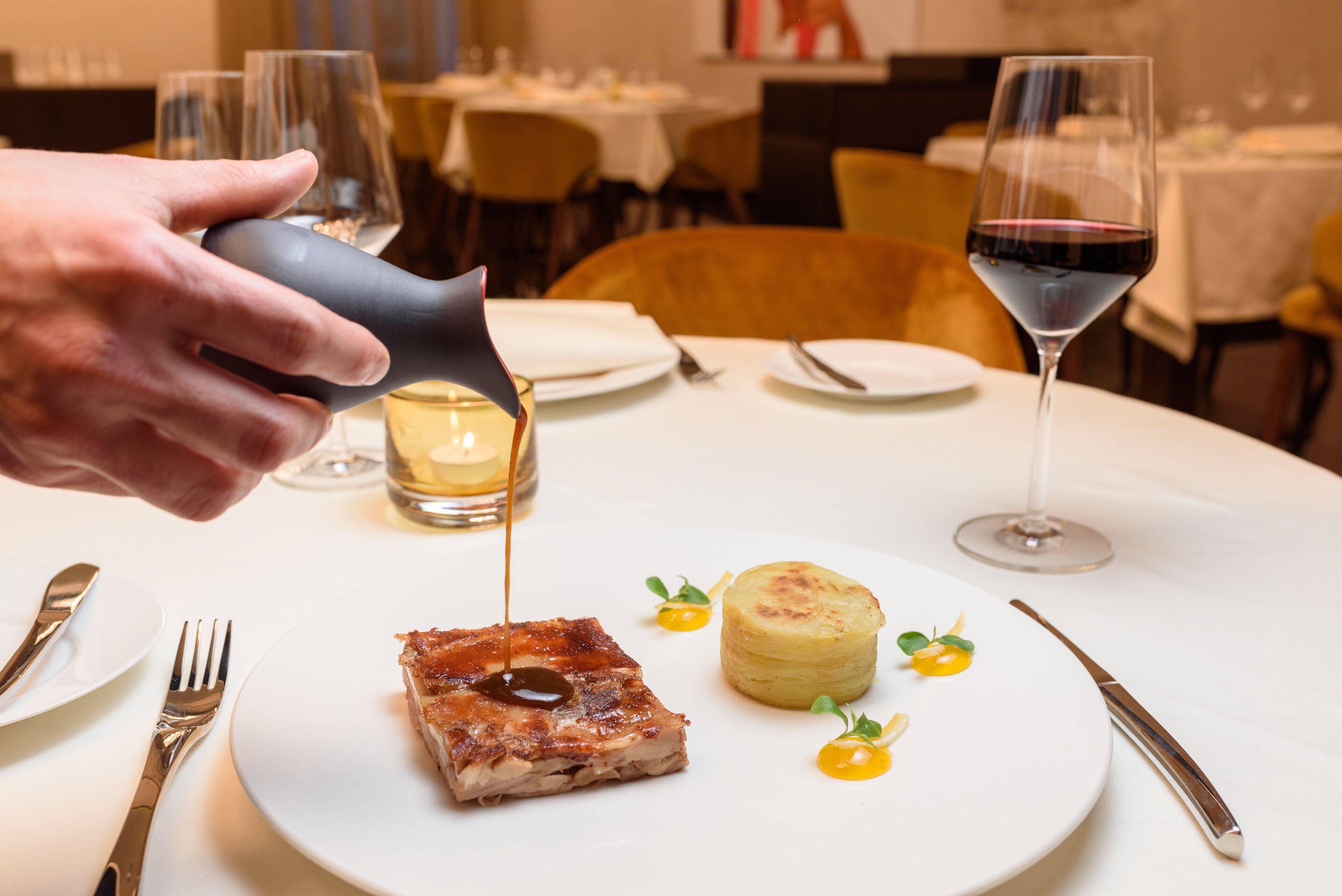 A dining table with a plate of food, a glass of red wine, and a hand pouring a drink.