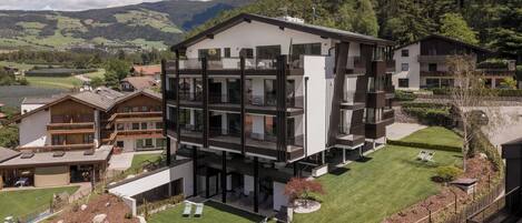 Un moderno edificio de varios pisos con balcones, un césped bien cuidado y montañas al fondo.