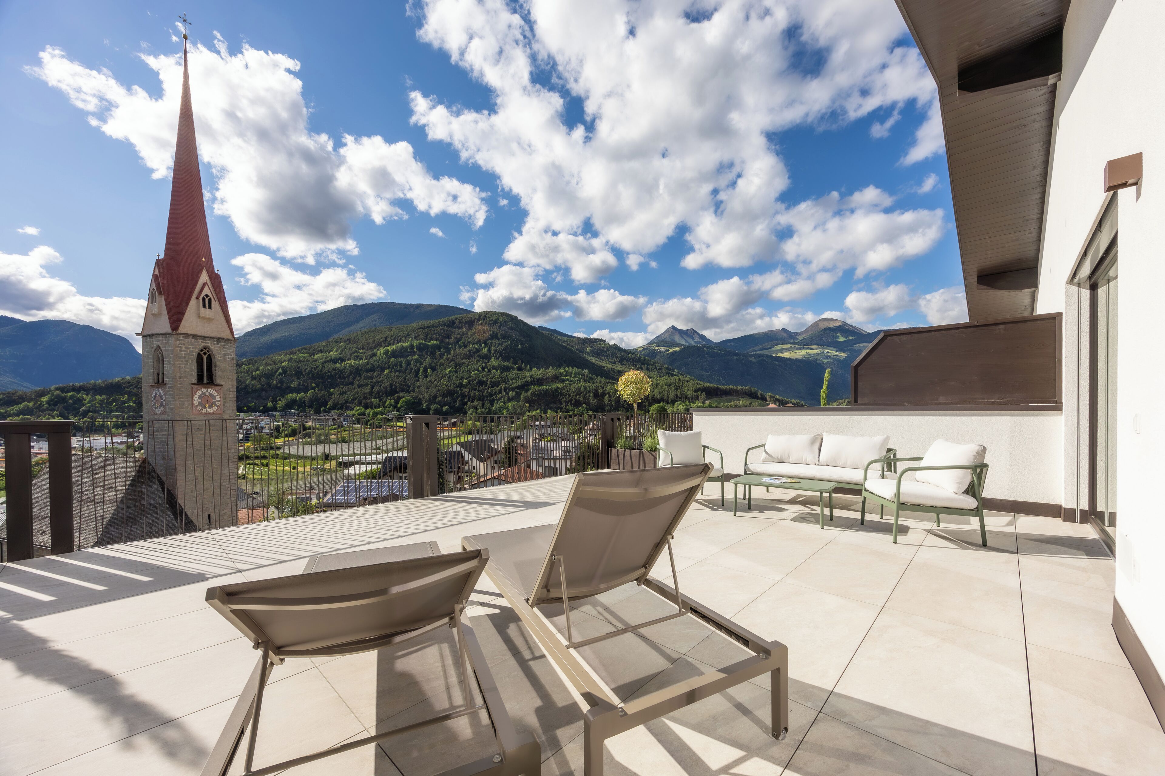 A terrace with lounge chairs, a clock tower, and a mountainous landscape.