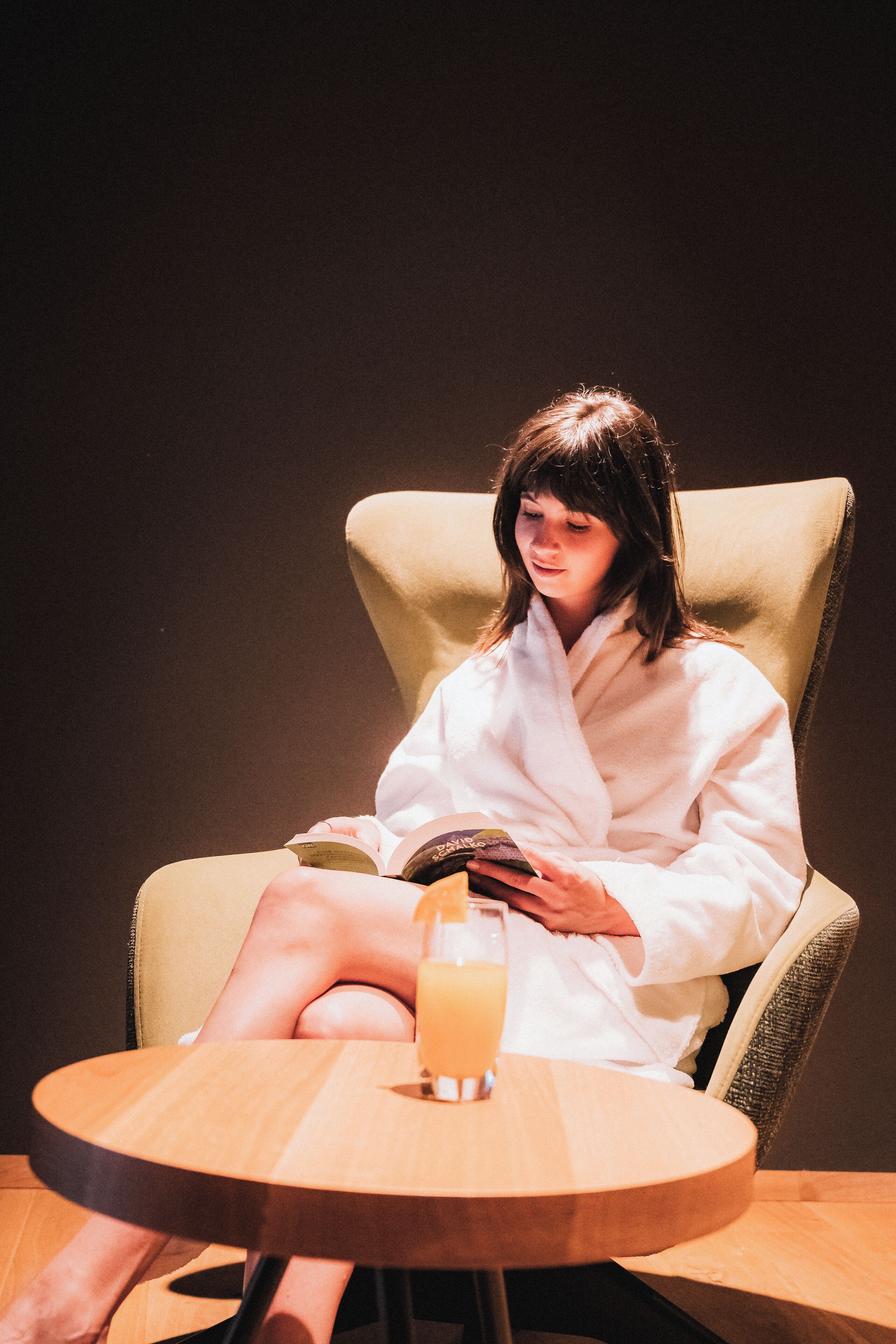 A person in a white robe sitting in a beige armchair with a round wooden table in front, holding a book and a glass of orange juice.