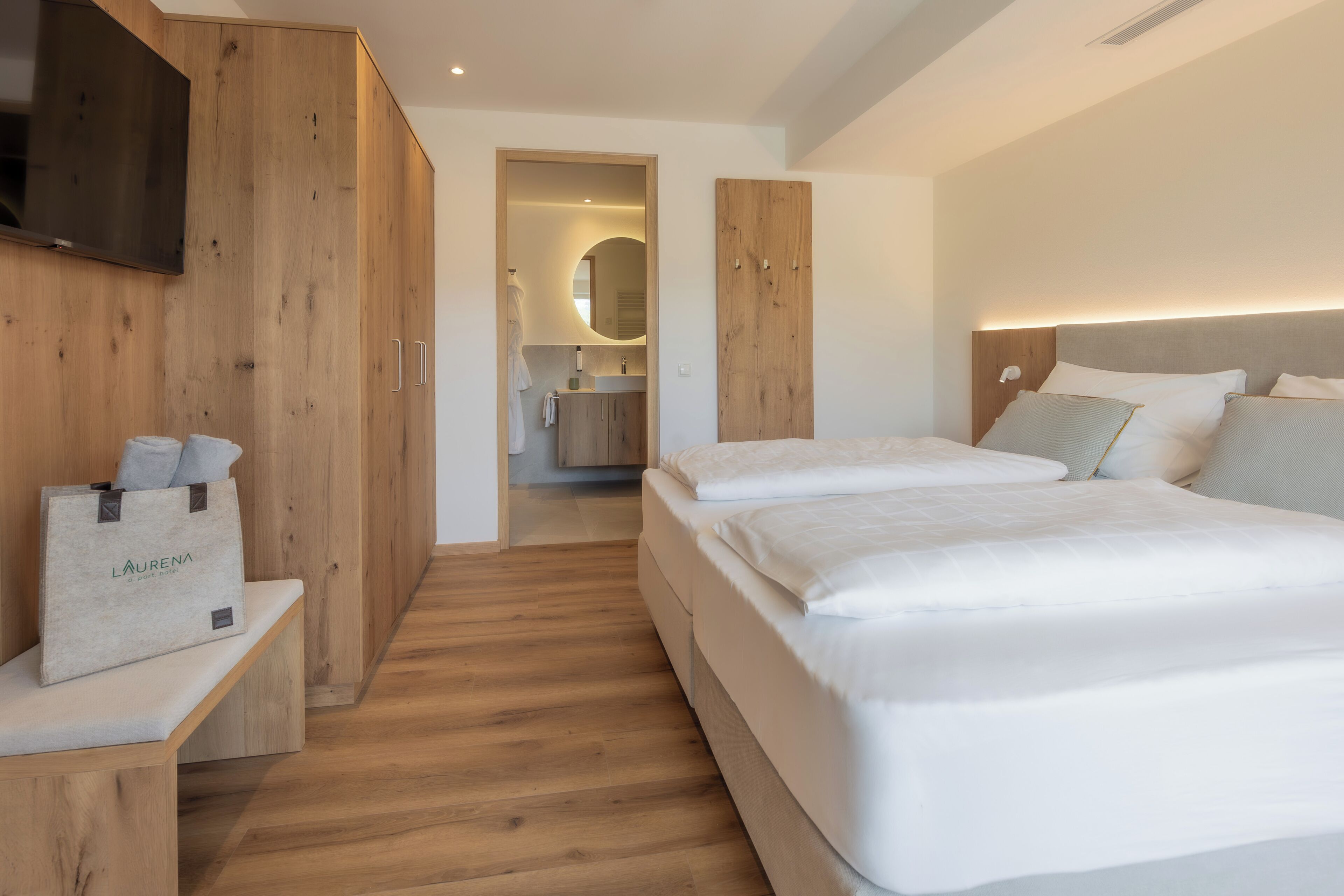 Ein modernes Hotelzimmer mit einem Holzschrank, einer Bank mit einer Tasche, einem großen Bett und einem durchgegangenen Tür erkennbaren Badezimmer.
