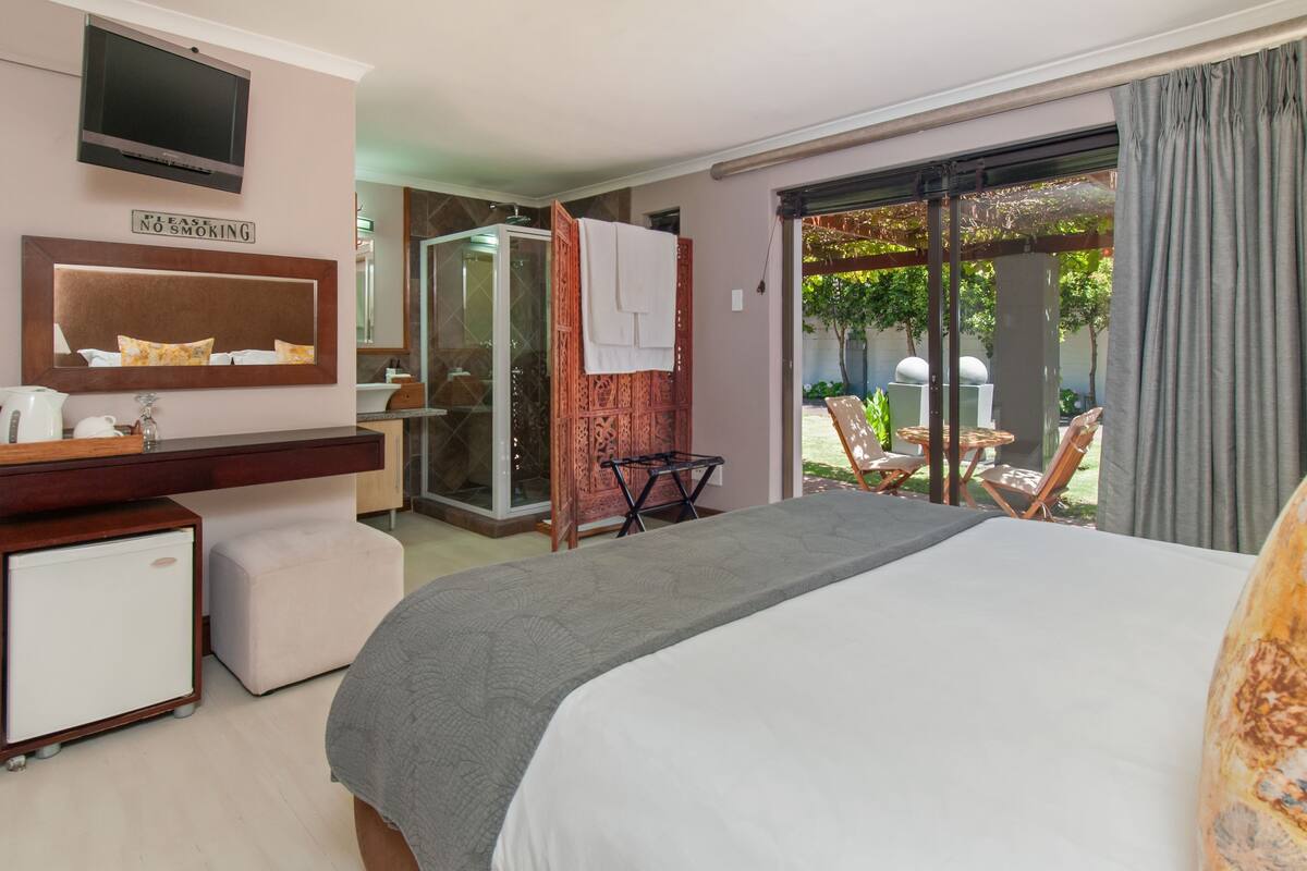 Ein Hotelzimmer mit Bett, Fernseher, Minikühlschrank, Spiegel, Dusche und einem Balkon mit Sitzgelegenheit.