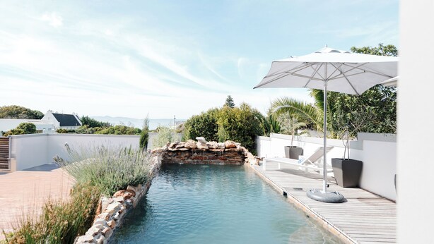 A clear pool with a wooden deck, surrounded by a white wall and greenery. - Whale Away Guest House (Hermanus)