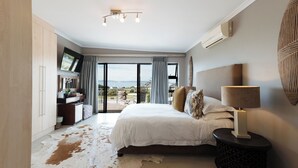 A bedroom with a large bed, a TV mounted on the wall, a nightstand with a lamp, and a view of the outdoors through large windows. - Whale Away Guest House (Hermanus)