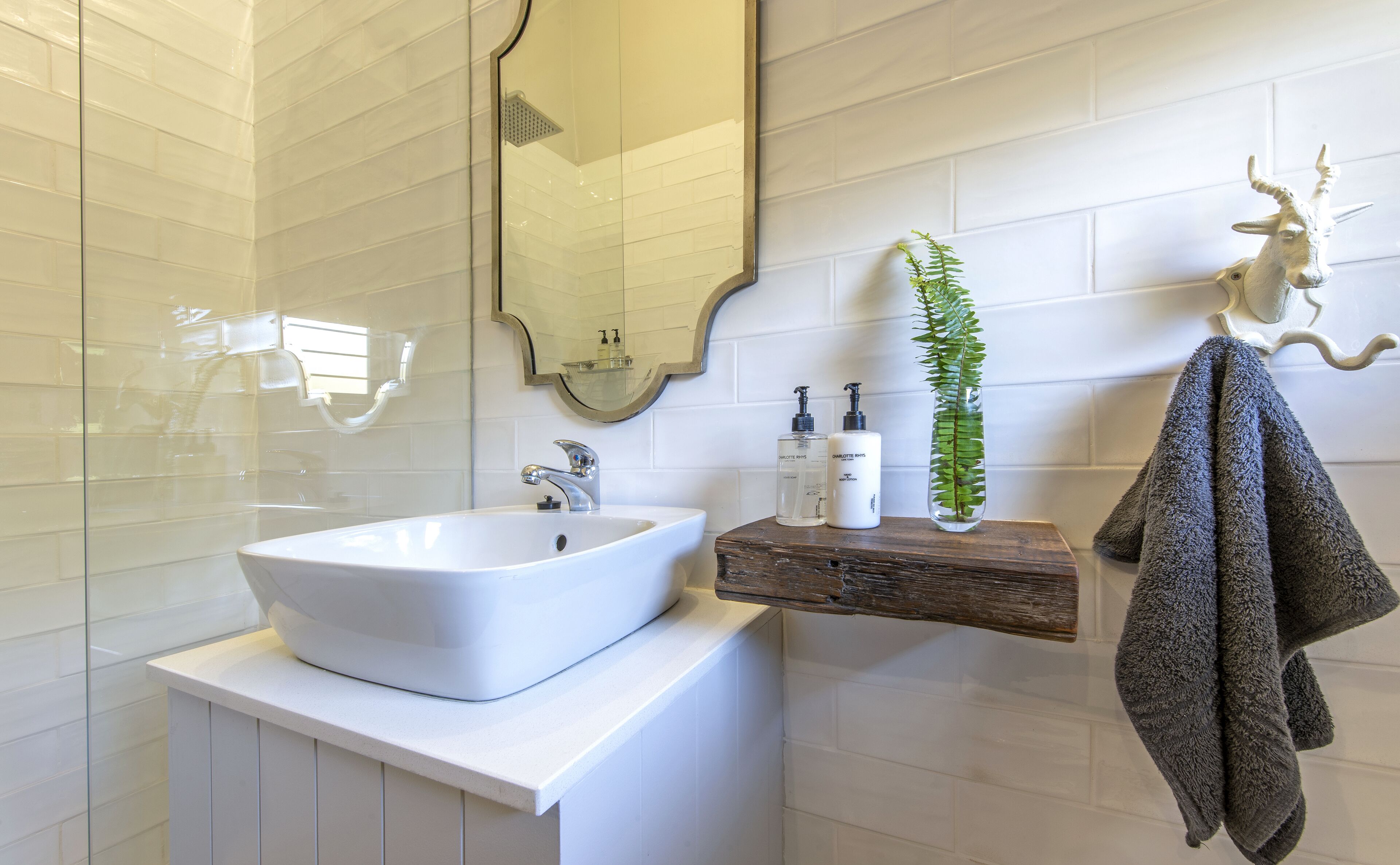 A bathroom with a white sink, a mirror, a wooden shelf with toiletries, a green plant, and a towel hanging on a hook.