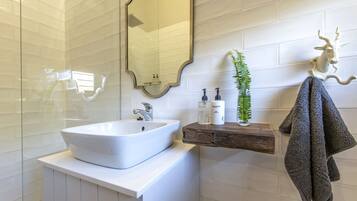 A bathroom with a white sink, a mirror, a wooden shelf with toiletries, a green plant, and a towel hanging on a hook.
