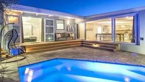 Un área de piscina moderna al aire libre con un porche de cristal, un banco de madera y una mesa de comedor con sillas.