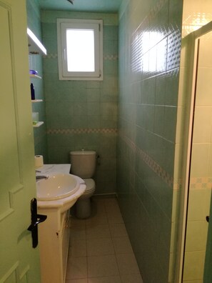A small bathroom with a toilet, sink, and a window above the toilet. Shelves are mounted on the wall, and the floor is tiled. - Bel Appart Au Cœur De La Marsa (La Marsa)