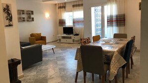 Un salon moderne avec un coin repas, comprenant un téléviseur à écran plat, des sièges confortables et une grande fenêtre avec des rideaux.