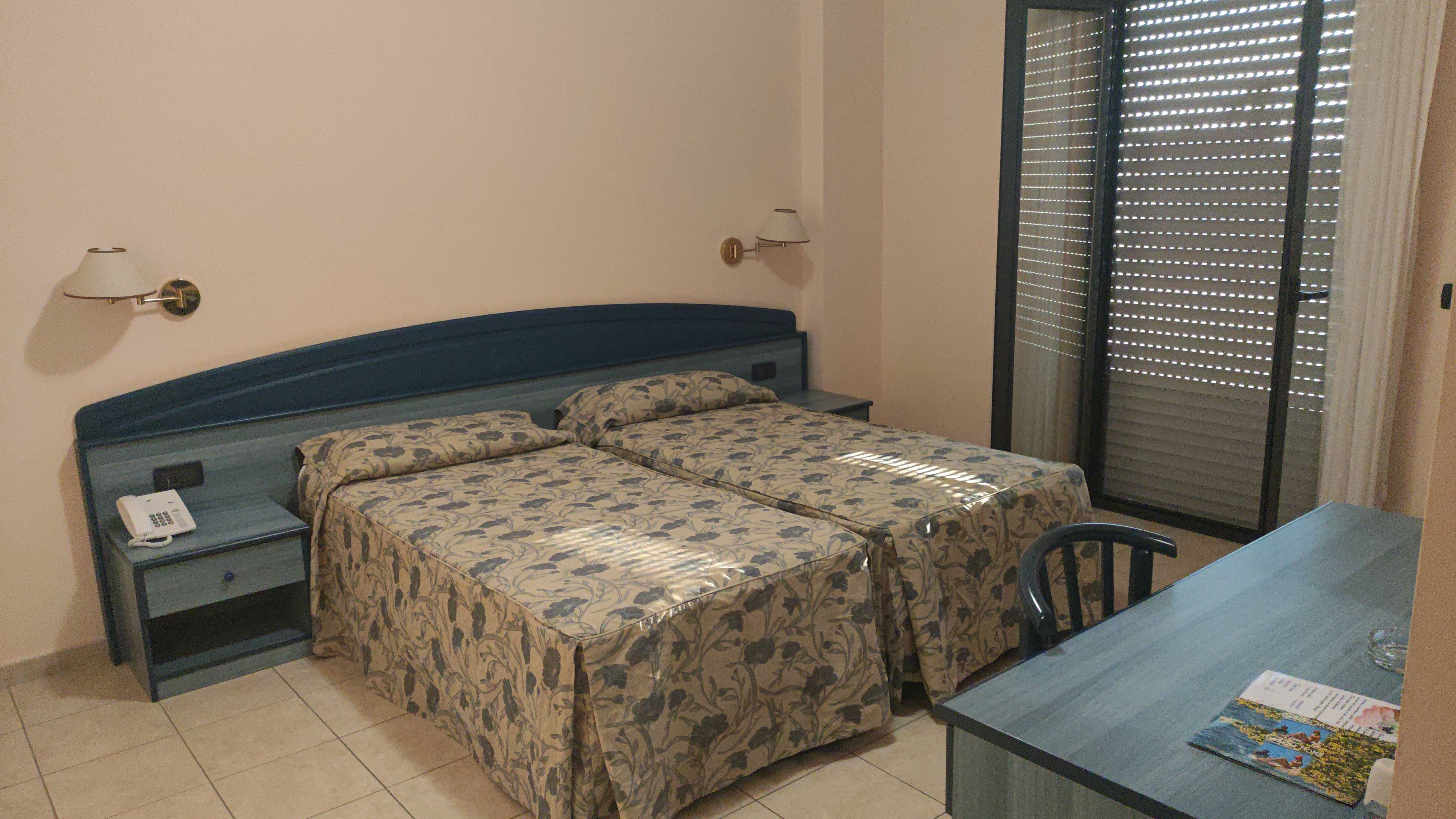 Una habitación de hotel con cama, mesitas de noche, escritorio y ventana con persianas.