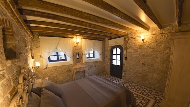 A bedroom with a stone wall, a wooden bed frame, a small bedside table, and a door with a window. - CavliHane 1885 (Ayvalik)